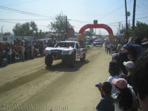 1990 Ford Bronco - white off-road trophy truck rolls along a dirt course lined with spectators, with a red inflatable arch in the background.