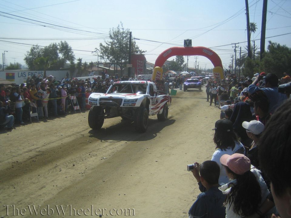 White off-road trophy truck on dirt course with spectators and inflatable arch