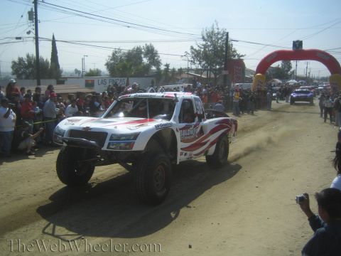 1990 Ford Bronco - white off-road trophy truck with red striping and Baldwin Motorsports decals drives along a dirt course before a crowd.