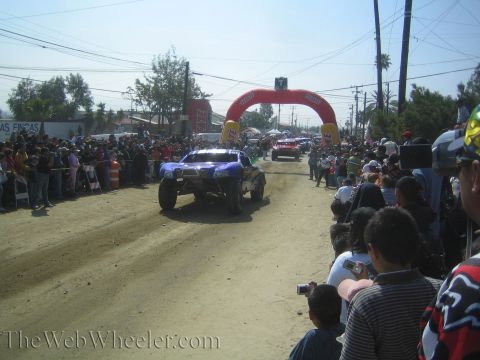1990 Ford Bronco - blue off-road trophy truck kicks up dust racing down a dirt road toward a red inflatable arch with spectators lining the route.