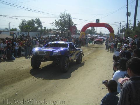 1990 Ford Bronco - blue off-road trophy truck rolls down a dusty dirt course as a crowd watches near a red and yellow inflatable race arch.