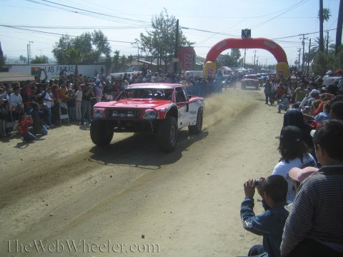 1990 Ford Bronco - red Ford off-road trophy truck catches air on a dusty dirt course as spectators watch and photograph from the side.