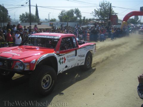 1990 Ford Bronco - red and white off-road trophy truck with Mexicana Logistics decals drives down a dusty course as a crowd watches.