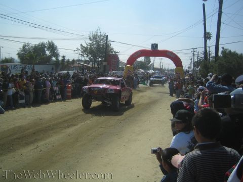 1990 Ford Bronco - red off-road trophy truck speeds down a dusty dirt road toward a red inflatable arch with dense crowds on both sides.