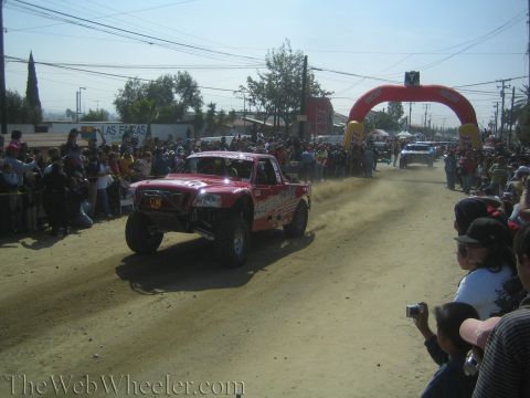 1990 Ford Bronco - red off-road trophy truck jumps on a dusty dirt course toward a red inflatable arch as spectators watch and film.