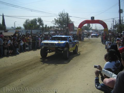1990 Ford Bronco - blue and yellow off-road trophy truck drives down a dusty course toward a red inflatable arch as crowds watch from both sides.