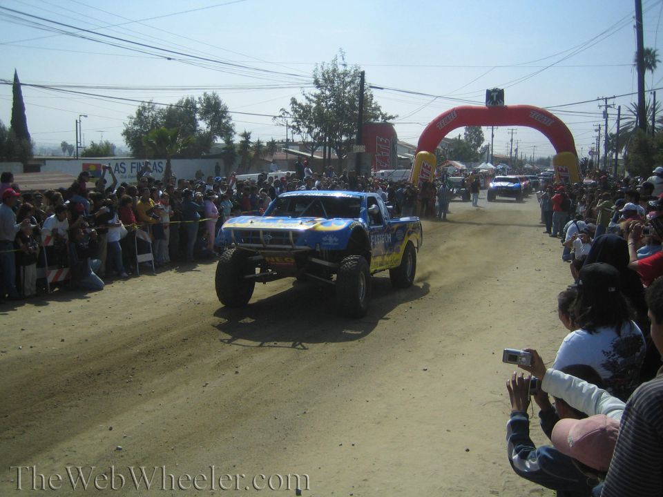 Blue and yellow off-road trophy truck on dirt course with spectators