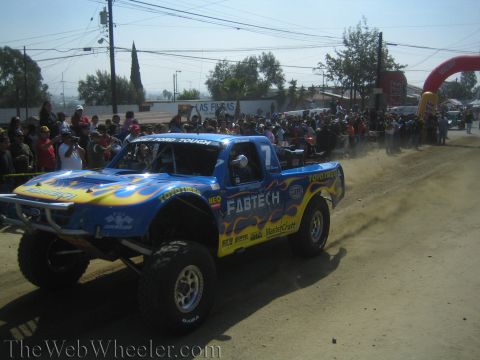 1990 Ford Bronco - blue off-road trophy truck with flame graphics and Fabtech and Toyo Tires decals, numbered 7, drives along a dusty course.