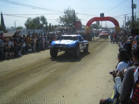 1990 Ford Bronco - blue off-road trophy truck rolls down a dusty dirt road toward a red inflatable arch with spectators lining the course.