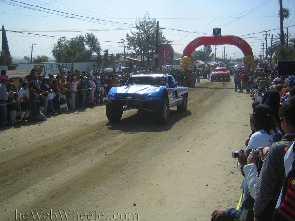 Blue off-road trophy truck driving down a dusty course toward inflatable arch