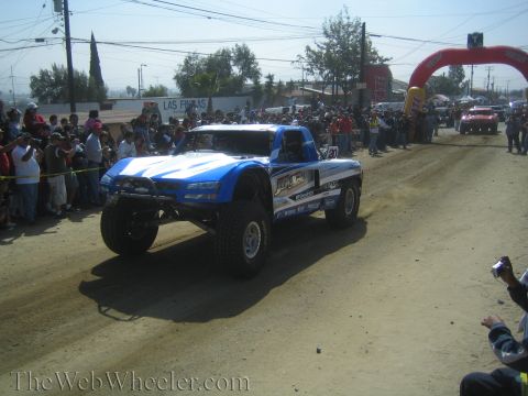 1990 Ford Bronco - blue and white off-road trophy truck numbered 1 drives along a dusty course before a crowd near a red inflatable arch.