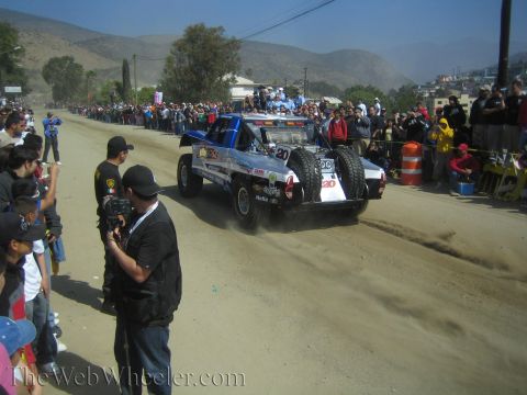 1990 Ford Bronco - blue off-road trophy truck numbered 20 drives down a dusty course backed by hills as a videographer and spectators watch.