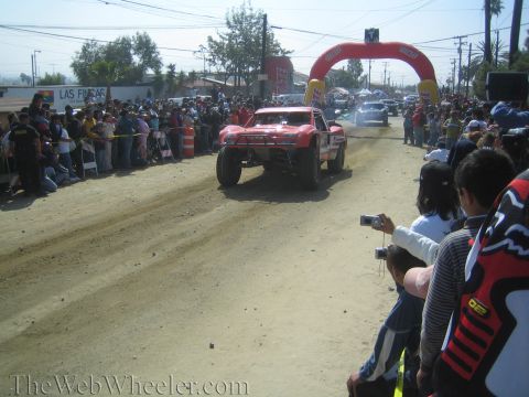 1990 Ford Bronco - red off-road trophy truck catches air on a dusty dirt course toward a red inflatable arch as spectators photograph it.