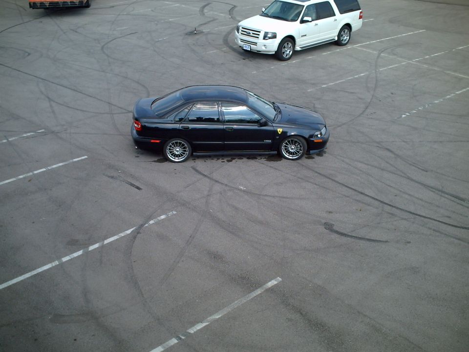 Dark sedan with silver alloy wheels in a parking lot full of tire skid marks, white SUV behind.