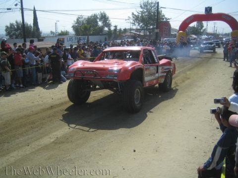 1990 Ford Bronco - red off-road trophy truck drives down a dusty course toward a red inflatable arch as spectators watch and take photos.