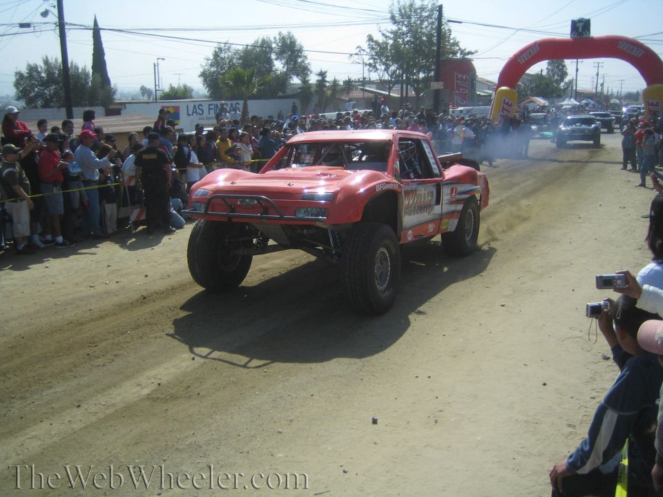 Red off-road trophy truck on a dusty course toward inflatable arch with crowd