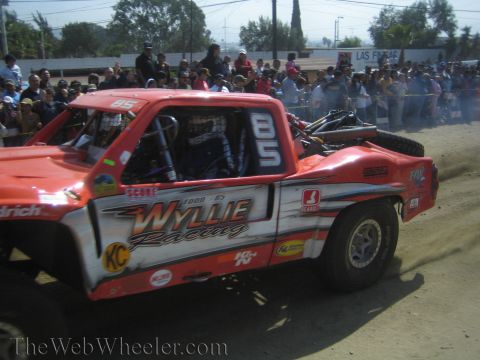 1990 Ford Bronco - orange off-road trophy truck with Wyllie Racing decals, numbered 85, drives along a dusty course as a crowd looks on.