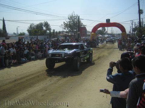 1990 Ford Bronco - black Monster-liveried off-road race truck kicks up dust on a dirt course lined with spectators and a yellow arch gate.