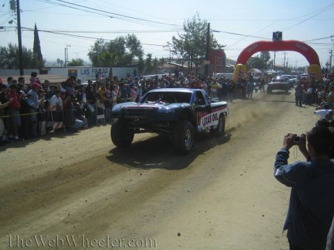 1990 Ford Bronco - off-road race truck wearing Lucas Oil sponsorship races down a dusty dirt course past a packed spectator crowd.