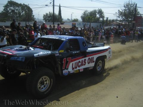 1990 Ford Bronco - blue and white number 17 Lucas Oil off-road trophy truck cuts across a dirt track in front of a dense crowd.