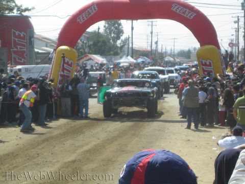 1990 Ford Bronco - off-road race truck drives under a red Tecate inflatable arch on a dirt course packed with spectators.