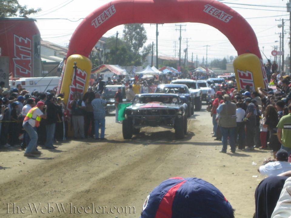 Off-road race truck passing under a red Tecate inflatable arch on a dirt course