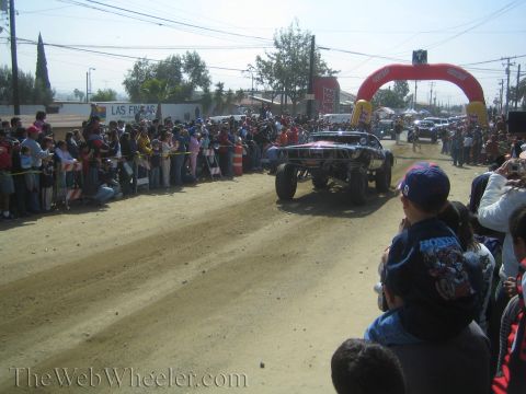 1990 Ford Bronco - black off-road race truck races down a dusty dirt course while spectators watch from behind the fence in the foreground.
