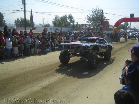 1990 Ford Bronco - black coupe-bodied off-road race truck speeds down a dirt course past a large crowd and an arch gate.