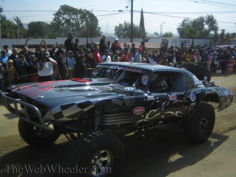 1990 Ford Bronco - black off-road race car with checkered-flag and flame graphics drives across a dirt course in front of cheering spectators.