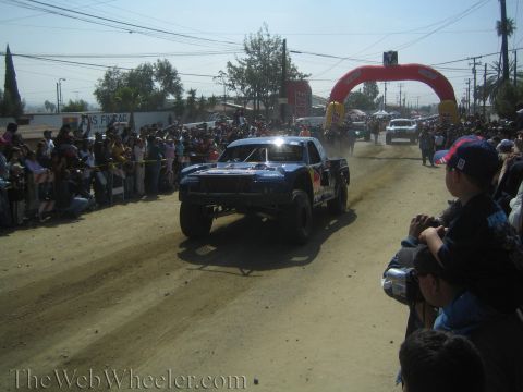 1990 Ford Bronco - dark blue Red Bull-sponsored off-road race truck races down a dusty dirt course lined with spectators.