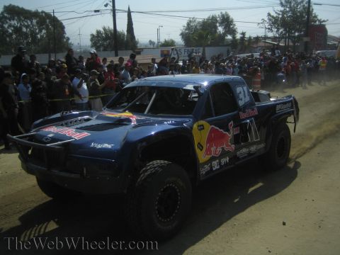 1990 Ford Bronco - dark blue Red Bull-liveried off-road trophy truck drives along a dirt road past a crowd of spectators.