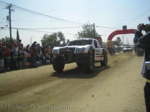 1990 Ford Bronco - white off-road race truck kicks up dust racing down a dirt course toward a red arch gate and spectators.