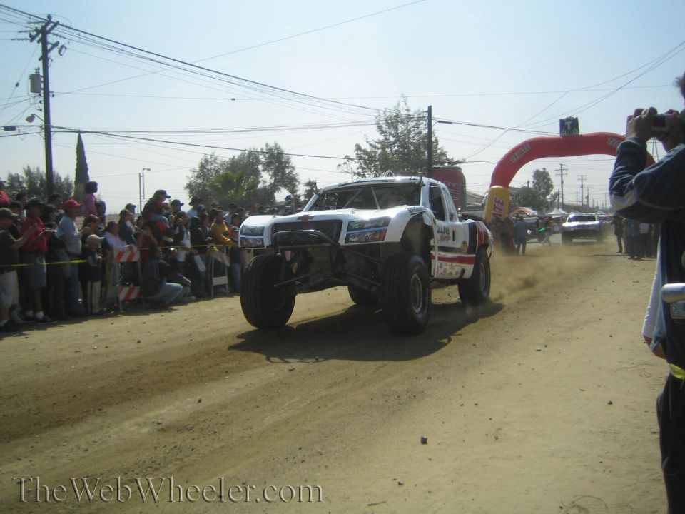 White off-road race truck speeding down a dusty dirt course past a crowd