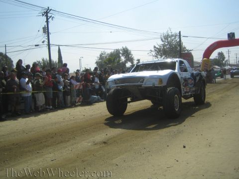 1990 Ford Bronco - white off-road race truck catches air on a dusty dirt course lined with a crowd and a red arch gate.