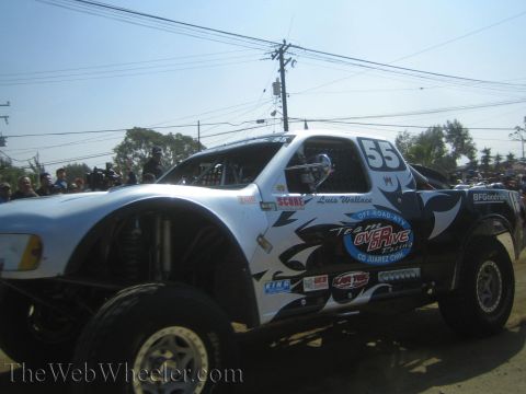 1990 Ford Bronco - white and black number 55 off-road race truck with tribal graphics drives along a dusty dirt course.