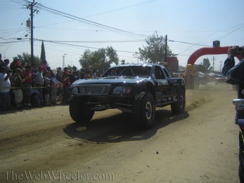 1990 Ford Bronco - black off-road race truck speeds down a dusty dirt course toward a red arch gate with spectators on both sides.