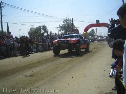 1990 Ford Bronco - orange off-road race truck races down a dirt course while spectators line the fence and an arch gate stands beyond.