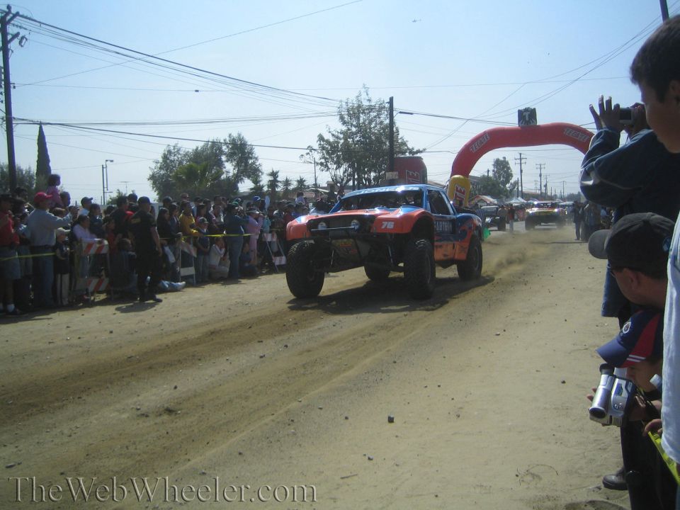 Orange off-road race truck racing on a dirt course past spectators