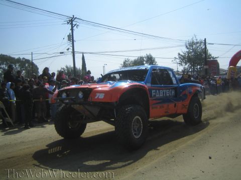 1990 Ford Bronco - orange and blue number 76 Fabtech off-road race truck drives down a dirt course in front of a crowd.