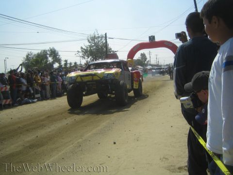1990 Ford Bronco - yellow off-road race truck kicks up dust on a dirt course lined with spectators near a red arch gate.