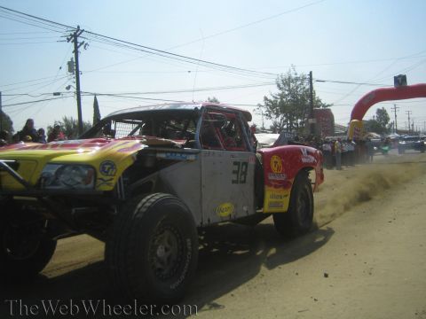 1990 Ford Bronco - Yellow and white number 38 off-road race trucks race side by side down a dusty dirt course past spectators.