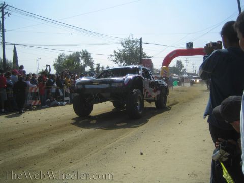 1990 Ford Bronco - silver and red off-road race truck races down a dusty dirt course toward a red arch gate with spectators looking on.
