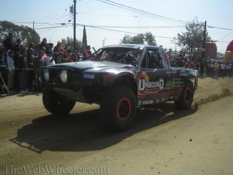1990 Ford Bronco - black off-road race truck wearing Unbound Energy livery and red wheels races down a dirt course past a crowd.
