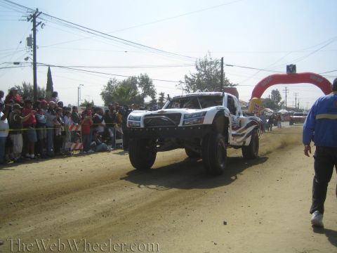 1990 Ford Bronco - white off-road race truck races down a dusty dirt course toward a red arch gate with crowds lining the fence.