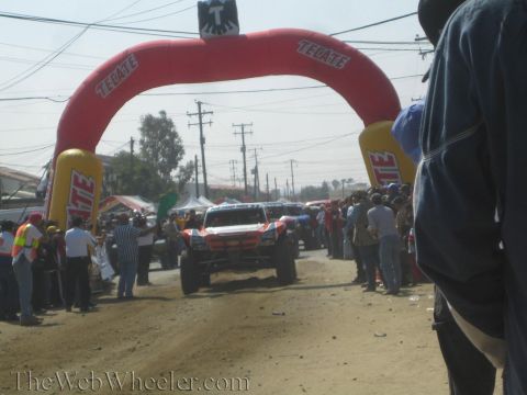 1990 Ford Bronco - Off-road race trucks gather under a red Tecate inflatable start arch on a dirt course flanked by crowds and tents.