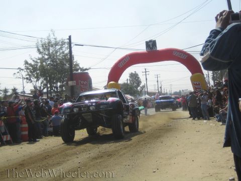 1990 Ford Bronco - Off-road race trucks kick up dust racing beneath a red Tecate inflatable arch on a crowded dirt course.