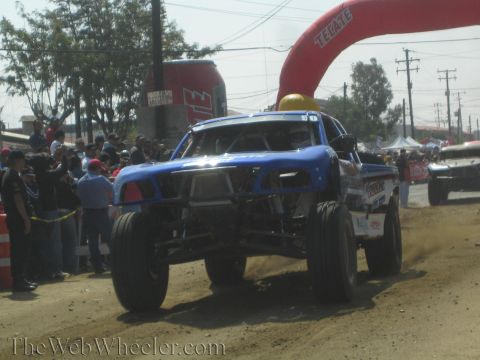 1990 Ford Bronco - blue off-road race truck races down a dusty dirt course near a red Tecate inflatable arch with spectators alongside.