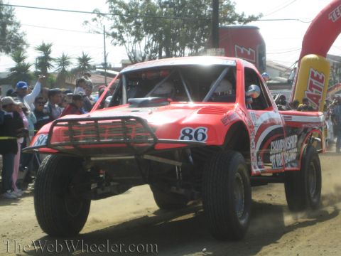 1990 Ford Bronco - red number 86 off-road race truck with a brush guard drives across a dirt course past a crowd and a Tecate arch.