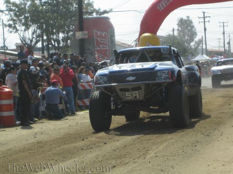 1990 Ford Bronco - blue No. 58 trophy truck with a Chevrolet bowtie launches off a dirt jump past spectators at an off-road race.