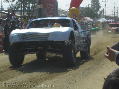 1990 Ford Bronco - silver No. 77 off-road race truck throws up a rooster tail of dirt as it races past spectators under a red inflatable arch.